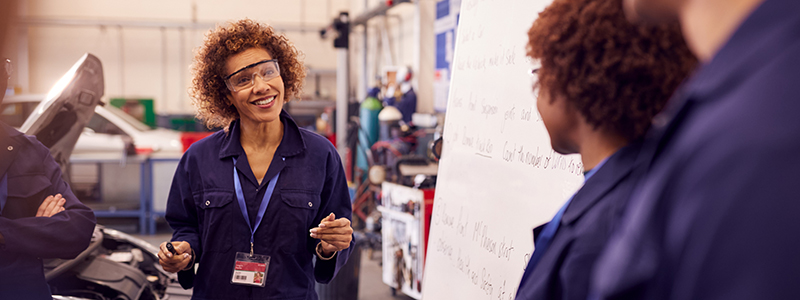 A smiling woman in safety glasses stands in front of a whiteboard looking towards a student